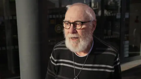 A man wearing a black jumper and silver necklace and black glasses. He is stood in front of automatic doors in a shop. 