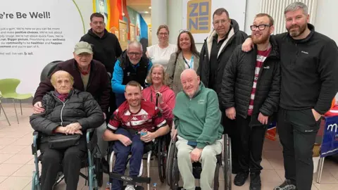 Jordan Burns A group of people gather in a bright, open indoor space with tiled flooring and white walls. Several individuals are seated in wheelchairs, while others stand around listening to someone speaking. Colourful balloons and a sign reading “ZONE” are visible on the wall.