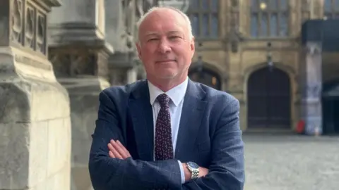BBC/Michael Keohan A man with short, white hair and a dark suit. He has his arms crossed.