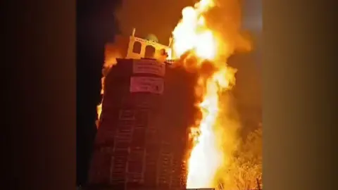 Social media A bonfire is on fire. An effigy of a model that looks like a mosque is on top with banners. It's night. The bonfire is leaning to the side.