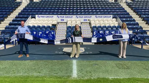 BBC/James Mountford A man and two women holding the long navy blue and white striped scarf on the pitch at PNE