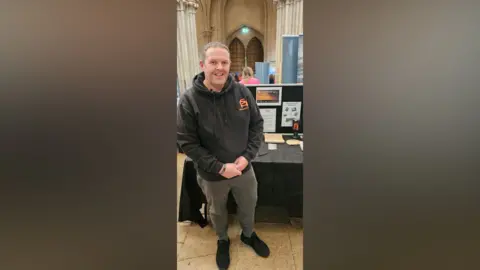 Handout Shane Green smiles at the camera as he stands in front of a black table. He is wearing a black hooded jumper, grey trousers and black shoes.