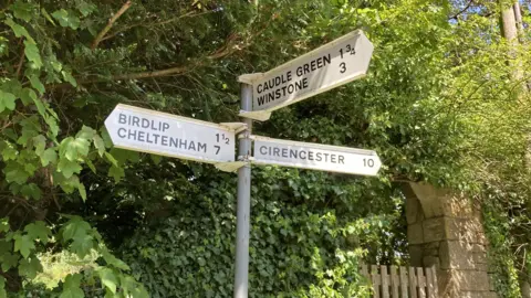 A photo showing a road sign with white finger post signs pointing in different directions. In the background are green trees and gated wall