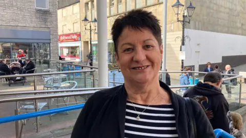 A woman with short, dark hair wearing a black and white stripy top underneath black jacket and standing outside 