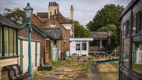 The HRE Group A heritage-style railway station platform lined with traditional brick buildings and vintage green-painted lamp posts. A dark railway carriage occupies the right-hand side of the image, while a wooden bench and station structures are visible on the left. The platform surface is weathered, with patches of dry grass growing between paving stones. In the background, mature trees and a railway bridge frame the scene under a cloudy sky.