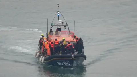 A RNLI boat, with blue hull and orange cabin, carries a group of people dressed in high-vis life jackets