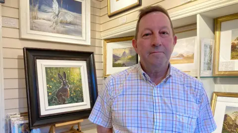 Tim Hardy has short brown hair. He is wearing a checked shirt. Behind him to the left there is a framed painting on an easel. It shows a rabbit or hare in a field with green flowers. There are other paintings in the background.