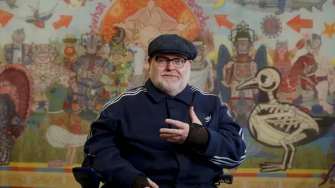 A man wearing a flat cap, a striped jogger style jacket and black glasses sits in front of a colourful wall or mural. He looks to be sitting in a wheelchair.