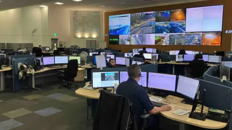 Sam Read/BBC The National Highways control room in South Mimms has a grid of cameras on the wall showing the major A roads and motorways in the East. There are workers sitting at screens taking calls and map of the roads is on a wall as well.