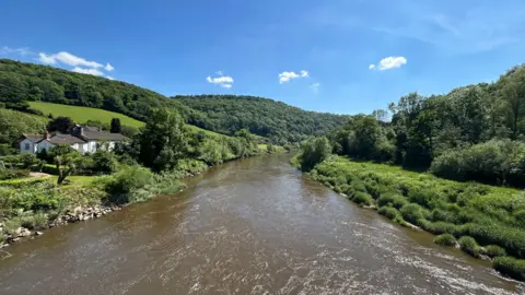 A view of the River Wye in Herefordshire flowing through countryside under a blue sky, with hills and trees on either side and houses at the foot of the hills on the rivers edge to the left.
