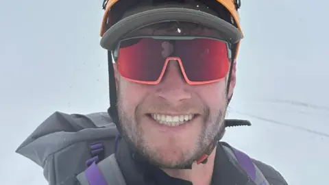 Henry Quinlan A man smiling in a selfie wearing sunglasses, a hard climbing hat and warm weather coat. The background is entirely white, with the snow on the ground blending into the white/grey sky.