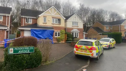 BBC A police car and a fire service vehicle are parked outside a property where the windows of the top floor are open and there is blue tarpaulin covering part of the lower frontage of the house.