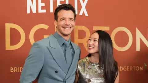 PA Media Luke Thompson smiles at the camera in a blue suit and tie and is stood beside Yerin Ha who wears a metallic dress and has long dark hair. They are stood against an orange screening backdrop