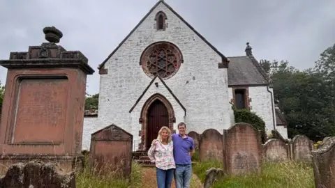 Kirsteen and Andrew Mitchell standing in front of whitewashed Applegarth church surrounded by gravestones 