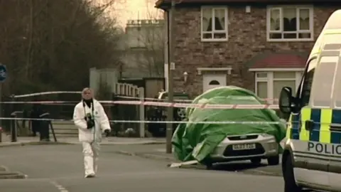 A blonde woman in white overalls walks in a residential road carrying a black camera around her neck. A liveried police van is in the foreground, while police tape surrounds a house and a silver Ford car covered with a green tarpaulin.