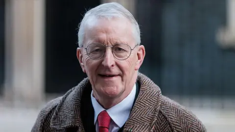 Hilary Benn has short grey hair, round metal glasses, a patterned coat, white shirt and red tie.