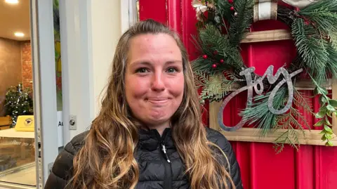 ELLEN KNIGHT/BBC A young woman looking into the camera and smiling slightly. She has long, wavy brown hair and is wearing a black puffer jacket. She's stood in front of a red door that has a green wreath on it, with the word 'Joy' written in silver letters. 