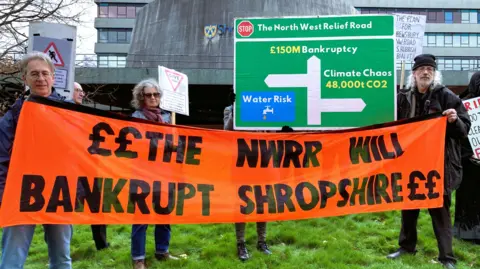 Two men and a woman holding up an orange banner than says "NWRR will bankrupt Shropshire". There is also a homemade green road sign behind them saying "Stop the North West Relief Road". In the background is an office block with bands of glass windows and cladding.