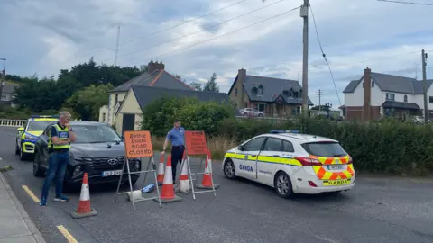 Scene of a road traffic accident. Two police cars are sandwiched between a large black car. Two officers are standing beside orange cones and road signs indicating the road is closed. 