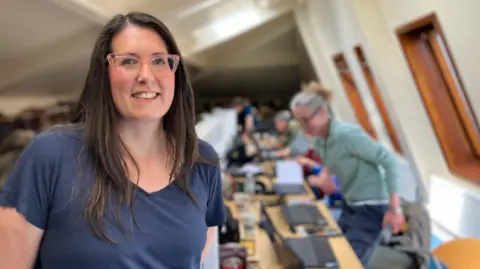 A woman with long dark hair, a dark blue t-shirt and pink-rimmed glasses smiles at the camera. Behind her are people using laptops