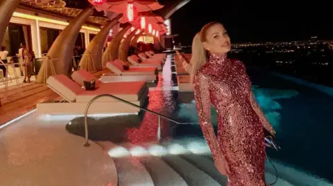 Handout Hofit Golan poses for a photo in front of a pool and sunbeds in the night time. She is wearing a glittery, pink, floor-length dress.