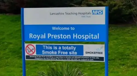 A blue and white sign reading 'Welcome to Royal Preston Hospital' stands amid greenery at the landscaped entrance to the hospital