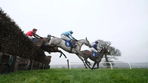 Getty Images Three riders are jumping a hurdle at the racecourse. The middle rider is wearing a blue jersey, the rider behind him a red one.