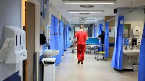 A man in red scrubs walks through a hospital ward, which is divided into treatment areas. You can see sinks, blue ward curtains and a dispenser on the wall for plastic gloves.