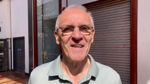 A grey-haired man wearing steel-rimmed glasses and a light grey polo shirt 