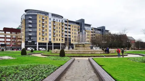 Queen's Court in Hull with yellow brickwork and grey steel cladding many of the flats have balconies and the penthouses are glass fronted. In the foreground is grass and a large fountain.