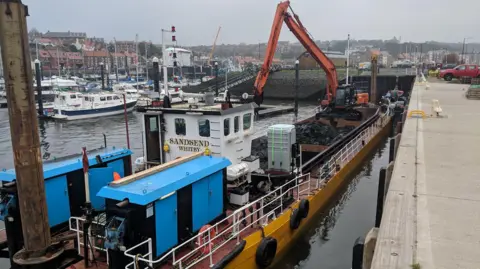 A dredging vessel alongside a harbour painted in a mix of blue, white and yellow, with the name Sandsend written on the white cabin.