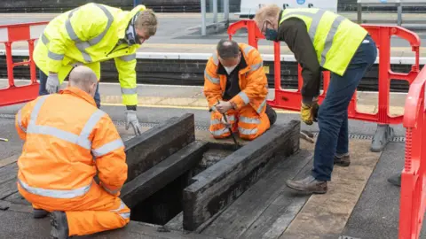 Sheaf and Porter Rivers Trus Four men with PPE inspecting the shaft underneath prior to installation of the light well on a railway platform.