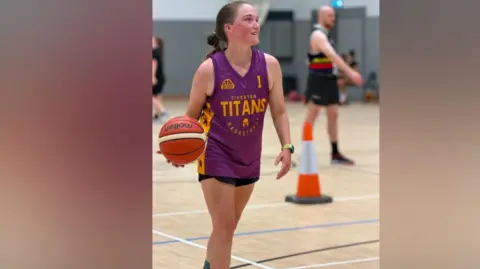 Tiverton Titans A young woman in a purple vest with the word Tiverton Titans in yellow is holding an orange basketball.