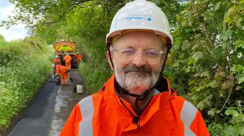 A man in an orange safety jacket with white hard hat. He has glasses and a grey beard. Behind him is a team of pothole repair workers with a lorry