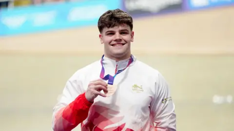 A man wearing a red and white tracksuit is seen smiling while holding a bronze medal. He is stood indoors in a brightly-lit sports arena.