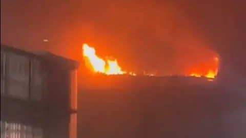 Jenna Tilley Orange flames on the roof of the building light up the night sky. Another building can be seen on the left.
