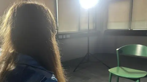 Susan is sitting to the left of the picture but you can only see her shoulders and her hair. In front of her is a green chair, and there is a large light in the corner. She is in an empty room with screens pulled down 