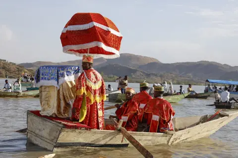 Anadolu via Getty Images A priest in a boat on the water is shaded from the sun by a large red umbrella. The boat is also carrying a replica of the ark covered in a gold cloth.