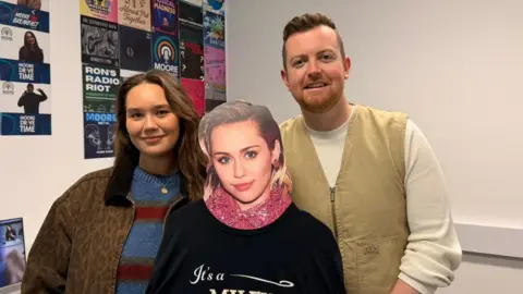 Darcy Davis and Jack Bostock Darcy wears a striped jumper and patterned jacket and has brown hair. She stands next to Jack who wears a white jumper and cargo vest and between them is a Miley Cyrus cutout 