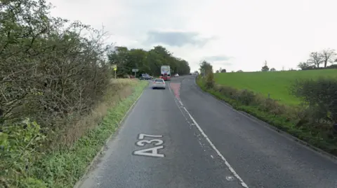 A road in a rural area shows a line of cars travelling north from the camera. On the left and right are grassy verges.