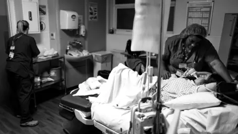 James Clifford Kent A wide black and white shot of a hospital delivery room where a midwife in scrubs leans over a patient on a bed, while another staff member works at a station in the background.