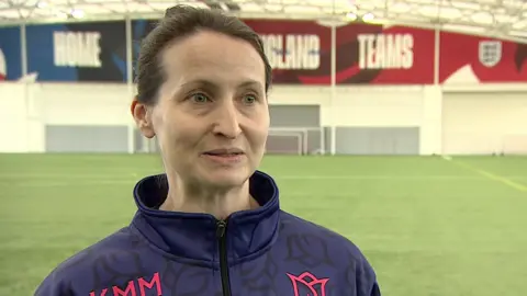 A women with brown hair tied up stood on a football pitch. She wears a blue top with some pink writing on it.