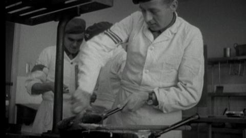 An army chef stirring something in a saucepan while two more chefs work behind him.