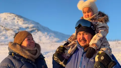 Angutimmarik Hansen wearing glasses and a beanie carrying a small child on his shoulders his wife standing next to him smiling