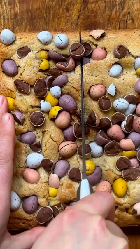 A hand holds a knife cutting a cake full of mini eggs