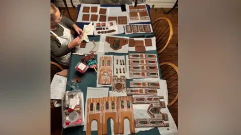 Julie Hamer The photo shows a large table completely covered with pieces of a miniature cathedral gingerbread construction project.
A person sits on the left side of the table working with a piping bag of white icing. Their face is not clearly visible.
The table is filled with neatly arranged gingerbread components in many shapes and sizes, each decorated with intricate white icing patterns.