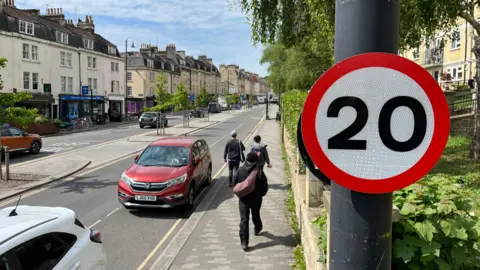 A 20mph sign on a lamp post along London Road in Bath, Somerset. Buildings can be seen either side of the road as well as parked and moving cars. A few people are walking along the pavement.