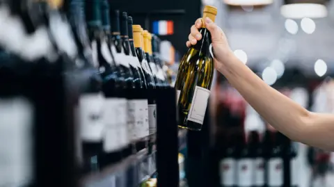 A woman grabs a bottle of wine from a shop shelf.