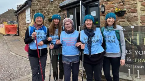 A group of five women stand together in front of stone buildings in hiking gear wearing vests reading "Mary's March".  They are all wearing knitted teal headbands, except the older woman in the centre who wears a pink headband. Her vest reads "Mary's march, walking for cancer aged 90."