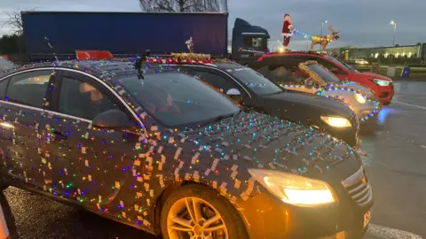 Four cars in a row with their headlights on. All the vehicles are decorated in festive lights. At the top of the photo is a model of a reindeer and Father Christmas. 
